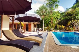 tropical gardens back a pool villa with blue swimming pool and sun loungers in foreground