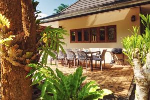 brick paved patio with tropical ferns around. Outdoor dining table and chairs with BBQ